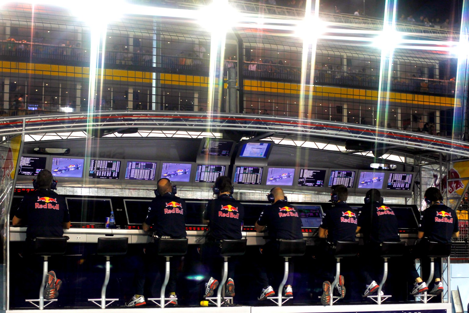 Ingenieros del equipo Red Bull con el logo del patrocinador trabajando frente a una pared de pantallas de datos en el muro de boxes de un Gran Premio de Fórmula 1