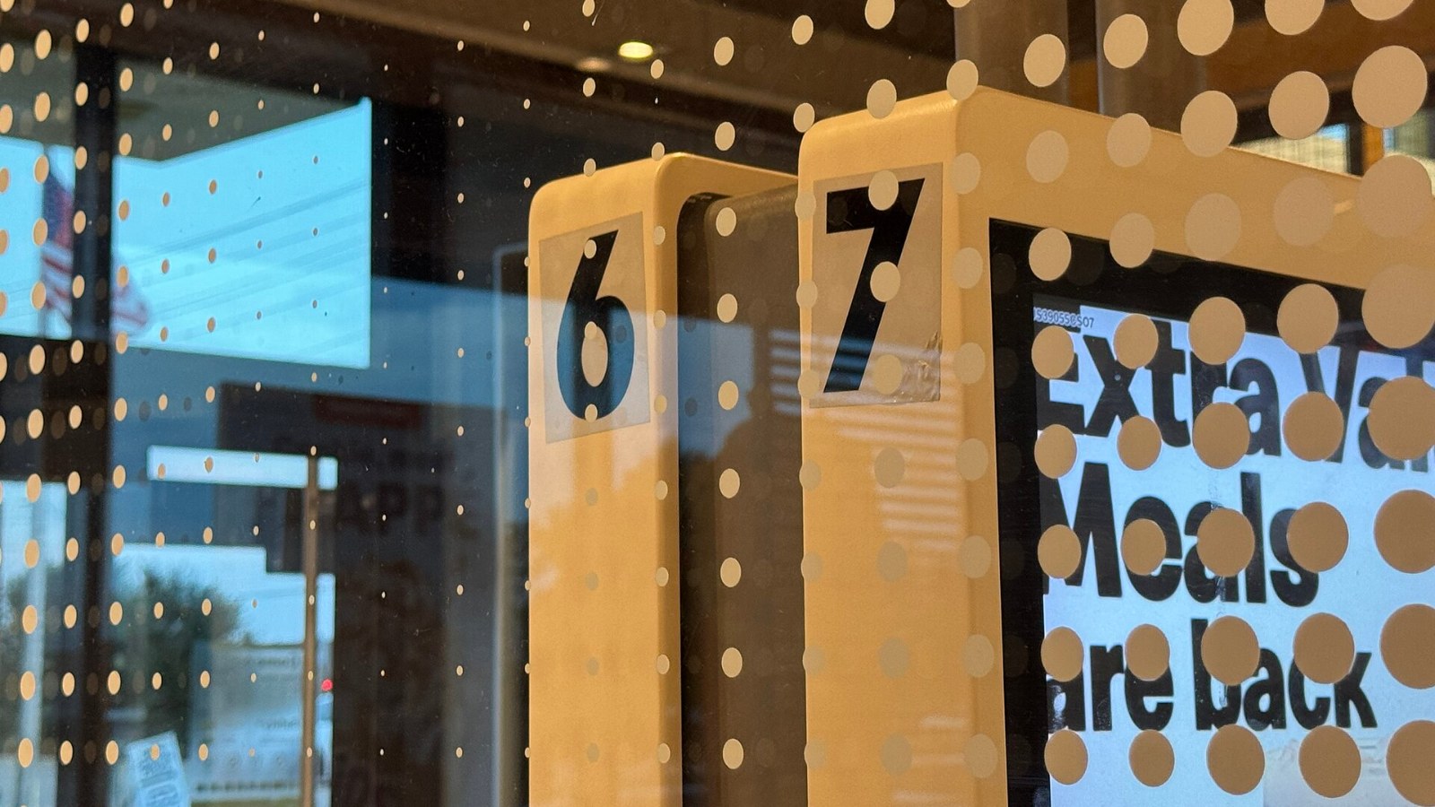 Dos kioscos de autopedido de McDonald’s con los números 6 y 7 y una pantalla que anuncia Extra Value Meals, vistos a través de un cristal