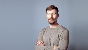 Retrato de MrBeast, Jimmy Donaldson, sonriendo con los brazos cruzados y jersey gris sobre fondo azul grisáceo, en 2026