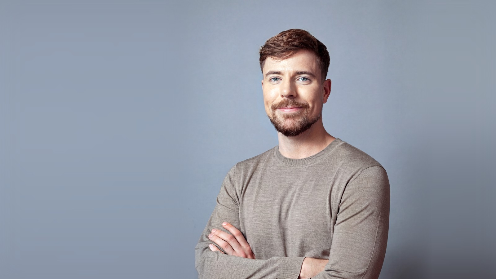 Retrato de MrBeast, Jimmy Donaldson, sonriendo con los brazos cruzados y jersey gris sobre fondo azul grisáceo, en 2026