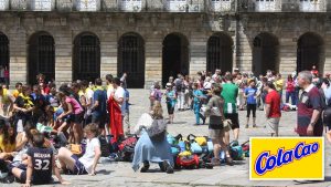 Plaza del Obradoiro de Santiago de Compostela llena de peregrinos sentados junto a sus mochilas, con el logotipo de ColaCao en una esquina