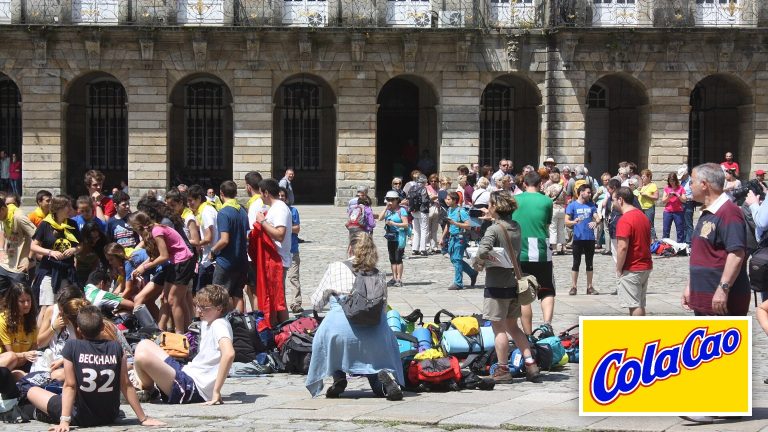 Plaza del Obradoiro de Santiago de Compostela llena de peregrinos sentados junto a sus mochilas, con el logotipo de ColaCao en una esquina