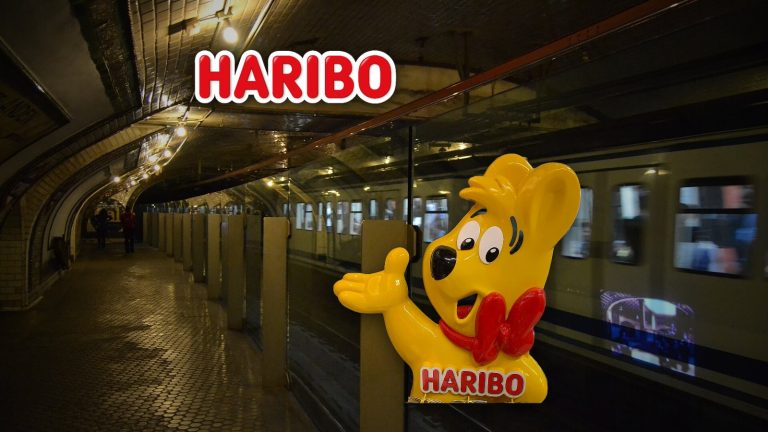 Logo de Haribo y figura del osito de oro de Haribo sobre el andén de la estación fantasma de Chamberí del Metro de Madrid, con un tren pasando