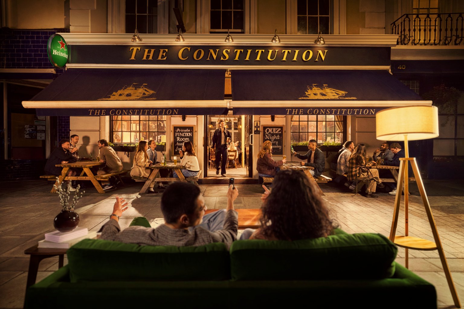 Imagen oficial de Heineken Pubscription: una pareja en un sofá con el mando a distancia frente a la fachada de un pub lleno de gente, como si el pub fuera la pantalla