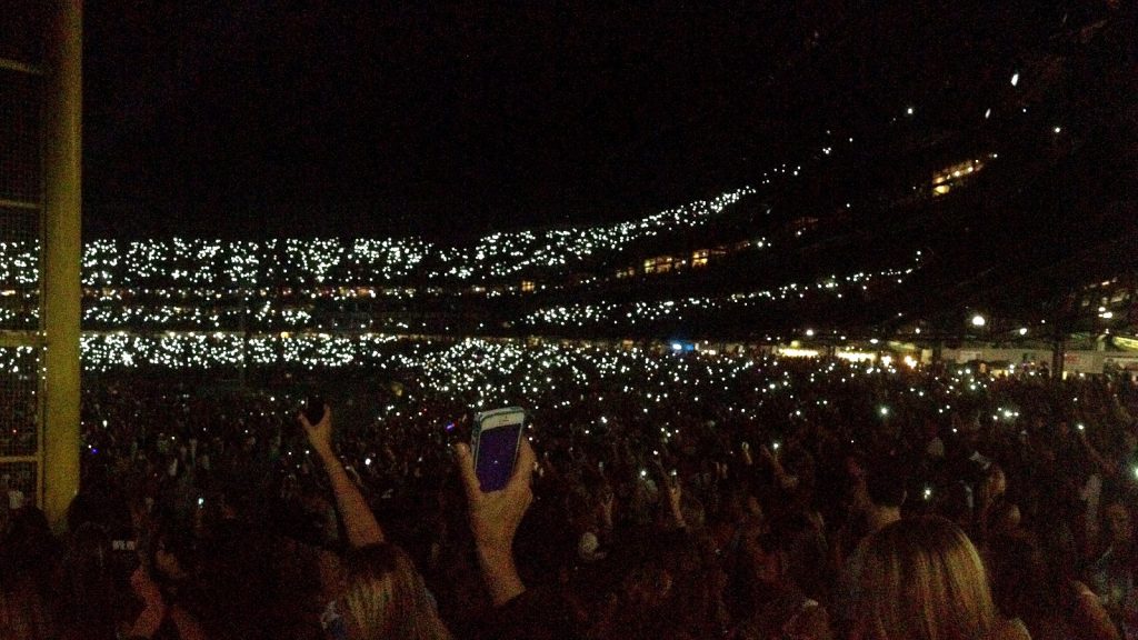 Miles de luces de móviles en las gradas de un estadio durante un concierto nocturno, con una mano en primer plano levantando un teléfono