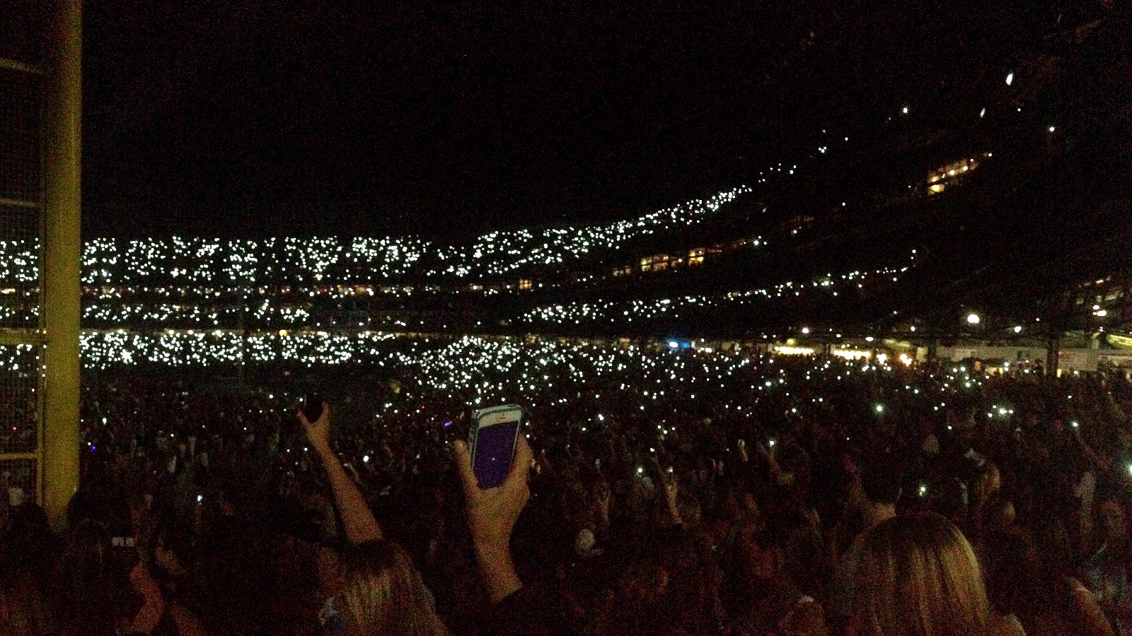 Miles de luces de móviles en las gradas de un estadio durante un concierto nocturno, con una mano en primer plano levantando un teléfono
