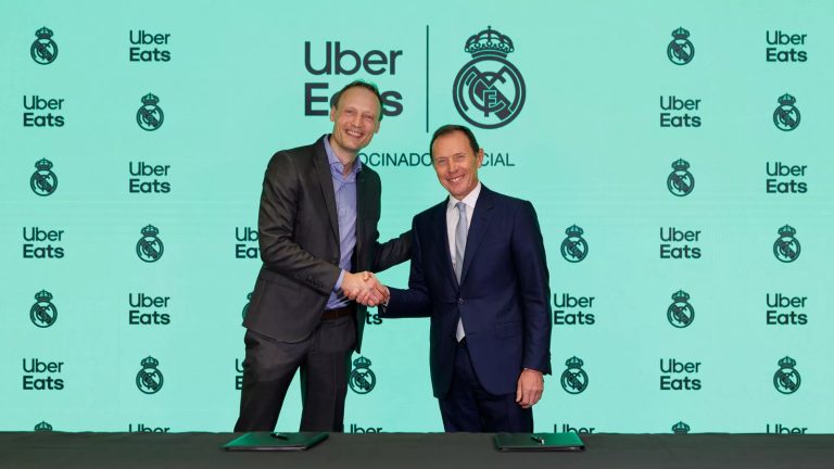 Representantes de Uber Eats y del Real Madrid se dan la mano ante un photocall con los logos de Uber Eats y el escudo del club tras firmar el patrocinio hasta 2029